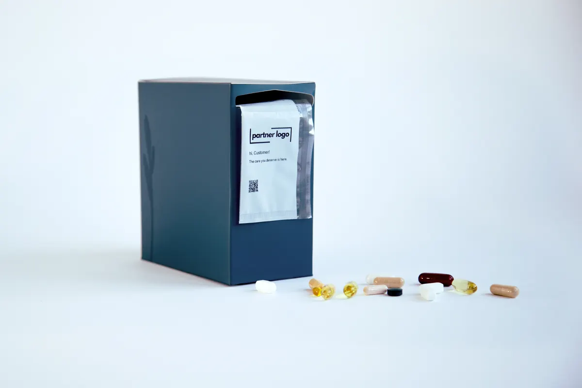 A dark blue dispenser box with a pack, printed with a practice's own logo, standing in the slot at its top, and loose capsules in front of it