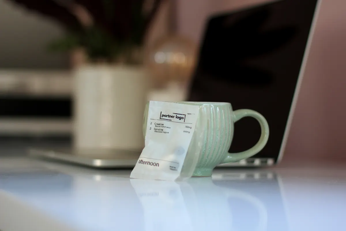 An afternoon pack, printed with a practice's own logo, resting against a cup of tea on a desk beside a laptop